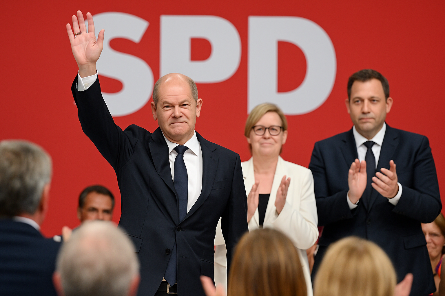 Olaf Scholz verabschiedet sich beim SPD-Parteitag 2025 in Berlin. Vor rotem Hintergrund mit großem SPD-Logo hebt er die Hand zum Gruß. Neben ihm stehen die neue SPD-Co-Vorsitzende Bärbel Bas und Lars Klingbeil, die beide applaudieren.