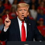 Das Bild zeigt den ehemaligen US-Präsidenten **Donald Trump** während einer öffentlichen Rede. Er steht hinter einem schwarzen Rednerpult, trägt einen **dunklen Anzug**, ein **weißes Hemd** und eine **rote Krawatte**. Mit erhobenem Zeigefinger und ernstem Gesichtsausdruck spricht er energisch in ein Mikrofon. Im Hintergrund ist eine unscharfe Menschenmenge zu erkennen, viele tragen **rote Mützen** und Kleidung – typisch für Trumps Wahlkampfveranstaltungen. Die Szene vermittelt Entschlossenheit und Dringlichkeit.