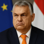 Porträt von Viktor Orbán, dem ungarischen Ministerpräsidenten, mit ernster Miene. Er trägt einen dunklen Anzug, ein weißes Hemd und eine orangefarbene Krawatte. Im Hintergrund sind unscharf die Flaggen der Europäischen Union und Ungarns zu erkennen.