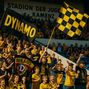Eine große Fangruppe von Dynamo Dresden schwenkt im Stadion der Jugend in Kamenz schwarz-gelbe Fahnen. Im Hintergrund jubeln Kinder im Dynamo-Trikot, die Tribüne ist voller Dynamo- und Oberlausitz-Fans.
