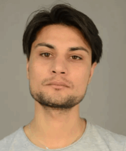 Fahndungsfoto eines Mannes mit dunklen Haaren, kurzem Vollbart und ernster Miene vor grauem Hintergrund. Auffällige Narbe am linken Unterarm und eine weitere markante Narbe an der linken Wange. Der Mann wird wegen Mordes gesucht