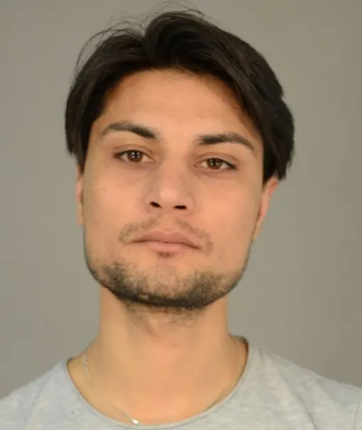 Fahndungsfoto eines Mannes mit dunklen Haaren, kurzem Vollbart und ernster Miene vor grauem Hintergrund. Auffällige Narbe am linken Unterarm und eine weitere markante Narbe an der linken Wange. Der Mann wird wegen Mordes gesucht