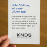 Eine Hand hält einen weißen Flyer mit der Aufschrift ‚Liebe Görlitzer, wir sagen Guten Tag!‘ und dem Logo von KNDS. Der Flyer liegt vor einem Holzuntergrund und wirkt wie eine Postwurfsendung an Görlitzer Haushalte.