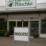 Das Foto zeigt den Eingang eines Gärtner-Pötschke-Geschäfts unter grauem, wolkenverhangenem Himmel. Im Vordergrund steht ein Metallgitter mit einem auffälligen weißen Schild, auf dem in schwarzen Großbuchstaben das Wort „INSOLVENZ“ steht. Dahinter erkennt man die Glasfront des Gebäudes mit dem bekannten grünen Firmenlogo von Gärtner Pötschke – einer stilisierten Gärtnerfigur mit Hut und Schürze. Durch die Scheiben sind Regale mit Pflanzen und Gartenartikeln zu sehen, doch der Eingangsbereich wirkt verlassen und ruhig. Die Szene vermittelt eine Mischung aus Stille und Unsicherheit.
