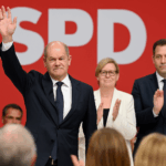 Olaf Scholz verabschiedet sich beim SPD-Parteitag 2025 in Berlin. Vor rotem Hintergrund mit großem SPD-Logo hebt er die Hand zum Gruß. Neben ihm stehen die neue SPD-Co-Vorsitzende Bärbel Bas und Lars Klingbeil, die beide applaudieren.