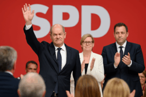 Olaf Scholz verabschiedet sich beim SPD-Parteitag 2025 in Berlin. Vor rotem Hintergrund mit großem SPD-Logo hebt er die Hand zum Gruß. Neben ihm stehen die neue SPD-Co-Vorsitzende Bärbel Bas und Lars Klingbeil, die beide applaudieren.